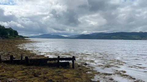 Loch Fyne at St Catherines - a muddy area leads down a gentle slop into a loch with mountains in the background on a cloudy day