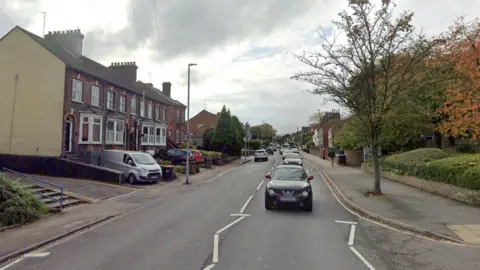 A view of West Street in Dunstable. The road is lined by pavements on each side, while the left hand side is lined with terraced houses. Other vehicles can be seen moving along the road.