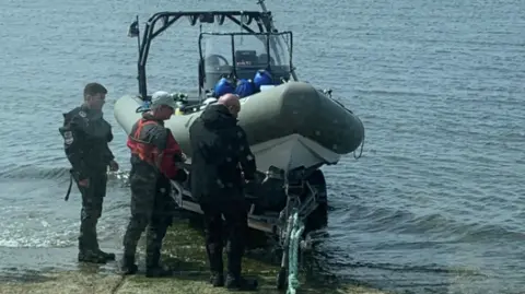 Clean Planet UK Three men dressed in diving gear are standing next to a ship which is being dragged out of the water by a blue rope.