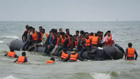 A crowded inflatable boat carrying migrants in choppy coastal waters, with several people in life jackets entering or surrounding the vessel.