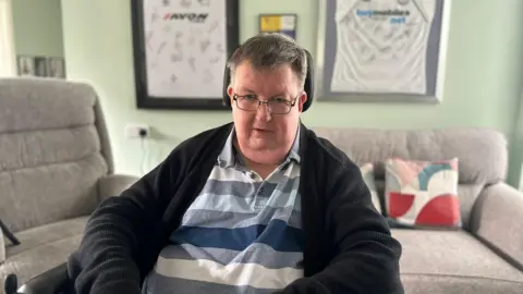 BBC A man with grey hair and glasses whilst being sat in a wheelchair inside his living room