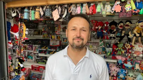 Sam Read/BBC A picture of a man, stood in front of his market stall which seels toys. He has a white polo shirt on and has a black goattee beard and short black hair.