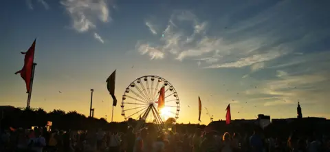 Shirley McInroy The sun slowly setting over a music festival crowd. There is a large ferris wheel and flags