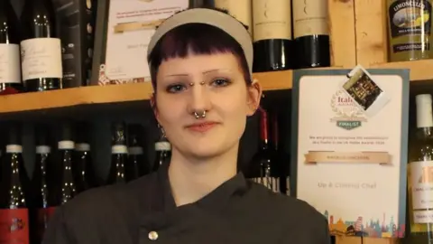 Buccelli's Evie Rei smiles as she poses in her chef uniform in front of her restuarant's shelves packed with wine bottles.