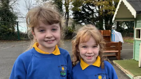 BBC Two girls stand in a school playground. They are identically dressed in a school uniform of a blue jumper with a school crest on and a yellow polo shirt underneath. The girl on the left is taller, with fair hair and the girl on the right has red curly hair. Both are posing and smiling for the camera. 