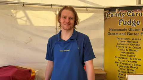 Liz Nice/BBC A man in a blue apron stands behind his stall, smiling. Behind him is a sign showing all the different fudge flavours available
