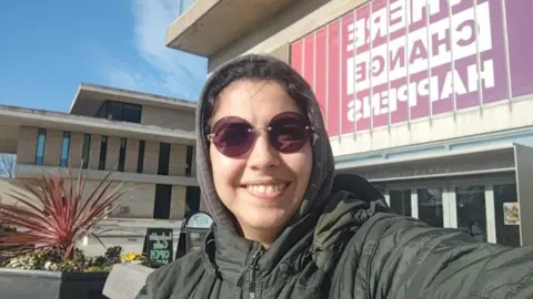 Family handout Vitória is in a khaki coat with a brown hood, and sunglasses, standing in front of brutalist 1960s concrete buildings at a university campus on a sunny day. The building nearest has large letters printed on a red-coloured backing.