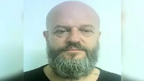 A bald man with a grey/ brown beard standing in front of a blue background wearing a black t-shirt.