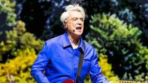 Getty Images David Byrne performs on stage. He plays a guitar and sings into a microphone that is strapped on to his head. He wears a blue jacket. 