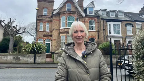 A woman with short white hair smiles as she stands in front of a striking, yellow-brick Victorian house with bay windows, gables and a tower. She is wearing a green coat and silver earrings.