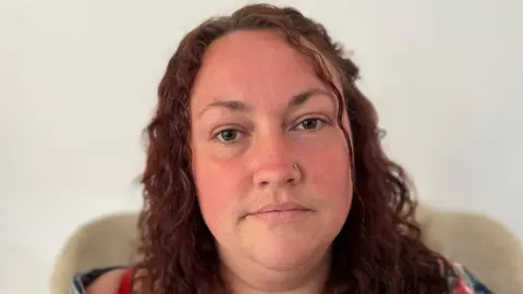 A woman with curly brown hair, brown eyes and a nose piercing. She is sitting on a chair against a white background.