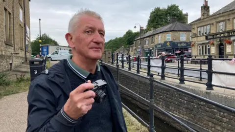 A man holding a camera in his right hand wearing a blue hood coat and standing next to a canal with black railings on either side.