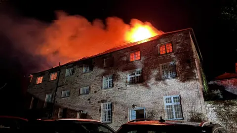 Torpoint Community Fire Station A three-storey stone-brick building is on fire at night with the majority of the flames on the third floor.