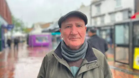 A man wearing a cap, grey scarf and green coat is looking at the camera. The background, which includes people on a walkway and buildings either side of it, is blurred.