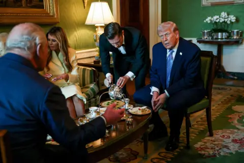 Suzanne Plunkett / PA Media The royal couple and the Trumps, all formally dressed, talk over tea in room with green walls as a server pours tea