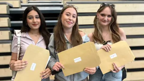 Sofia Raza, Shannon Cairns & Zornitsa Nacheva standing in front of wooden slat panels in a school hall. They are smiling and holding brown paper envelopes which contain their results