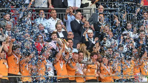 John Egan of Hull City in an orange playing shirt lifts the trophy after Hull City win the Championship play-off final with the players being showered in ticker-tape