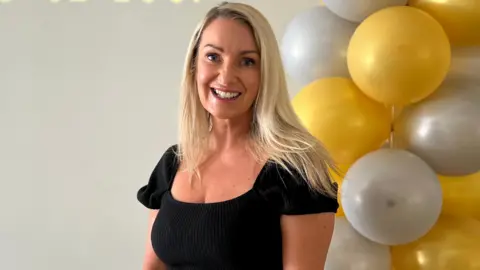 Tracy Stephenson smiling at the camera and stood next to a yellow and silver balloon arch. 