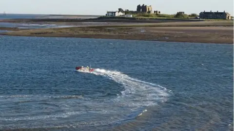 The lifeboat in the water travelling to the stranded pair. There is a trail of water behind the boat and the island can be seen in the background. 