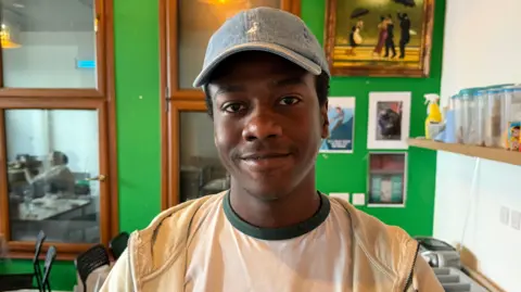 A young man wearing a blue baseball cap, white T-shirt and light hoodie stands smiling at the camera inside a community café or kitchen, with green walls, framed pictures and shelves in the background.