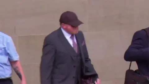 Robert Bush leaving court wearing a dark grey suit, white shirt and pink tie and pocket square he is wearing a dark baseball hat
