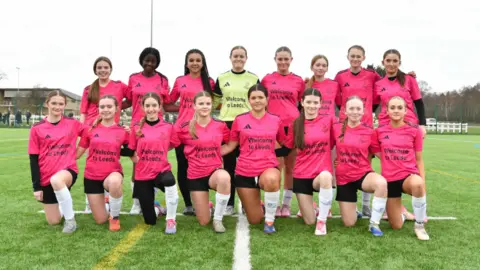 A group picture of girls on a football pitch wearing matching kits, which consist of pink t-shirts, black shorts and white socks. They are split into two rows with several stood up and the rest kneeling down in front. 