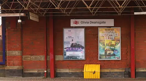 A train station sign reading 'Olivia Deansgate' hangs on a red brick station wall - posters advertising books can be seen on the station platform and a yellow grit box.