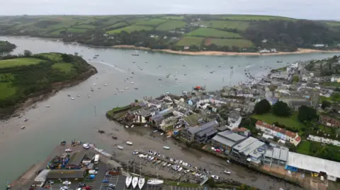 Aerial view of Salcombe in Devon. Boats are being stored in a car park. The estuary is full of boats. Green rolling hills can be seen in the distance. There are residential homes and businesses. 