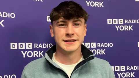 A young man stands in front of a purple BBC Radio York backdrop featuring repeated white text. He wears a light blue quarter-zip pullover over a white shirt.