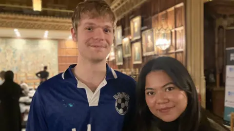 Shariqua Ahmed/BBC Will, a man wearing a blue jersey standing next to his partner Kia, a woman with short brown hair, smiling for the camera.