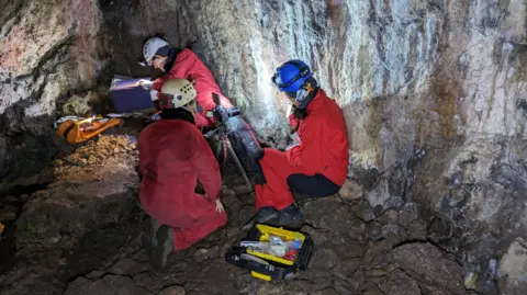 George Nash Three people in red boiler suits sit on a cave floor with the rock walls behind them. They wear helmets with torches on the front. A tool box lies on the ground by them. 