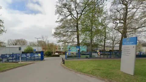 Google A school site, with a mainly grey and blue sign on the right including the words The Avon Valley School. Blue railings are on the left and right of the photo and there are trees on the right.