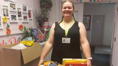 Debbie Taylor A woman is holding a basket of tins and cereals and is smiling at the camera. Behind her is an empty hallway and there are more boxes and bags of donations on her righthand side.