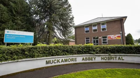 PA Media The exterior of Muckamore Abbey Hospital. There is a sign featuring the name of the hospital and it is also written on a low concrete wall. There is a hedge above this wall and a patch of grass in front. Behind it you can see two red brick buildings.