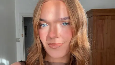 Roisin McMenamin Roisin McMenamin has long, brown, wavy hair and is wearing a green dress, standing beside a Christmas tree and a brick wall.