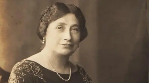 Black and white photo of a woman wearing a necklace and earrings. She has short hair