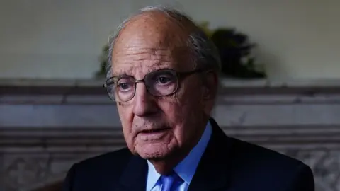 George Mitchell. He has grey hair at the sides, wearing glasses, a navy blazer, light blue shirt and blue tie. He is holding his hand out to the front. A marble fireplace is behind him.