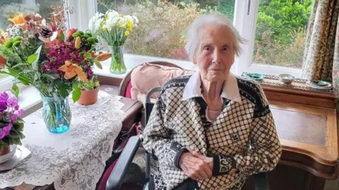 Family handout Sylvia Weatherhead sits on a chair in a house. She is wearing a black and white cardigan. Four vases and/or pots with flowers in them are on a table and windowsill next to her. 