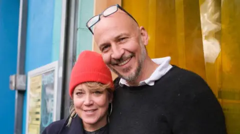 Kate Hodgkinson and Mark Collett. Mrs Hodgkinson has blonde hair which is poking out form under a red knitted hat. She is wearing large hoop earrings.  Mr Collett is bald with a grey beard. He has a pair of glasses resting on top of his head.