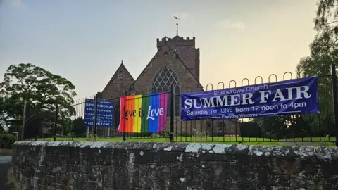 Ian Handley St Andrew's Church in Shifnal against the backdrop of an evening sky which is blue and soft yellow. There is a black metal fence with a large rainbow flag which reads "love is love"