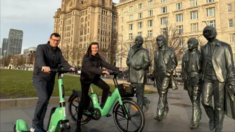 A man is on a green Bolt e-scooter and a woman is on a green e-bike. They are posing for the camera at Liverpool's waterfront in front of the Royal Liver Building and The Beatles statue