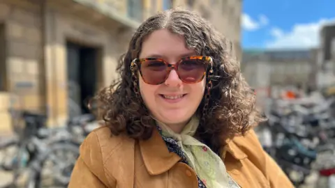 Head and shoulders image of Anna Ward. Anna has shoulder length curly dark hair, is wearing sunglasses and a camel jacket and lime green scarf.