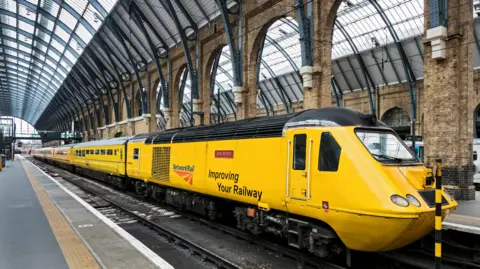 Network Rail A bright yellow train with a sloped front and multiple carriages on a railway inside a station. On the side of the train is a Network Rail logo and the words: "Improving Your Railway". The station is in the Victorian style with yellow-brick walls and pillars, steel roof supports and an arched glass roof.