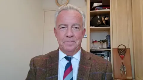 BBC News Sean Bell, wearing a brown tweed jacket and a red and blue striped tie, looks directly into the camera. He is sitting in a home office, and, in the background, various pieces of aviation memorabilia can be seen.