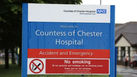 A close up of a blue, red and white sign saying: 'Welcome to Countess of Chester Hospital Accident and Emergency'. It is at the entrance to the A&E department with some of the hospital building and a couple of patients in the background. 
