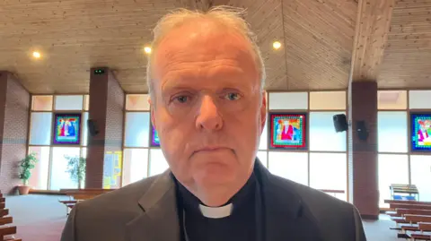 Fr Joe Gormley is standing in his church wearing a black suit and white dog collar.