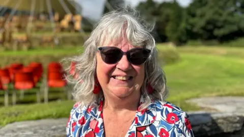 Vicki Gillings, a woman with grey hair and a fringe, she wears sunglasses and a flowery shirt, she stands in front of the Tynwald hill.