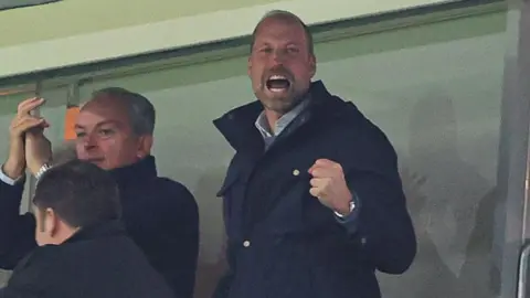 Prince William, wearing a blue coat, is cheering with his fist in the air at chest height