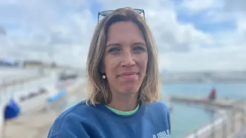 Nicola Murdoch who is wearing a blue jumper and has gold glitter on her face smiles at the camera while standing in front of Penzance's Jubilee Pool, which is blurred in the background.
