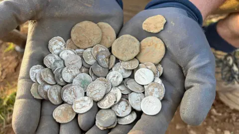 Michael Eakers/Brian Dixon A pair of dirty gloved hands hold a freshly unearthed hoard of worn silver and bronze Roman coins. 